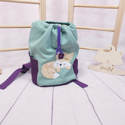 Geöffneter Kindergartenrucksack mit schlafendem Fuchsmädchen in Salbeigrün und dunkler Beere. Der Rucksack wird mit einer Kordel und einem Magnetknopf geschlossen